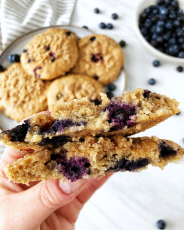 Blueberry muffin tops on a grey plate, a hand holding a muffin top and blueberries in a bowl