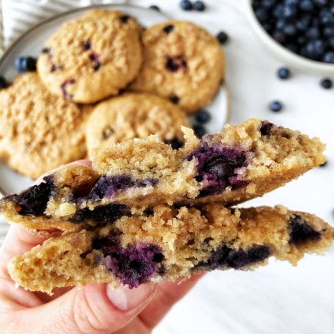 Blueberry muffin tops on a grey plate, a hand holding a muffin top and blueberries in a bowl