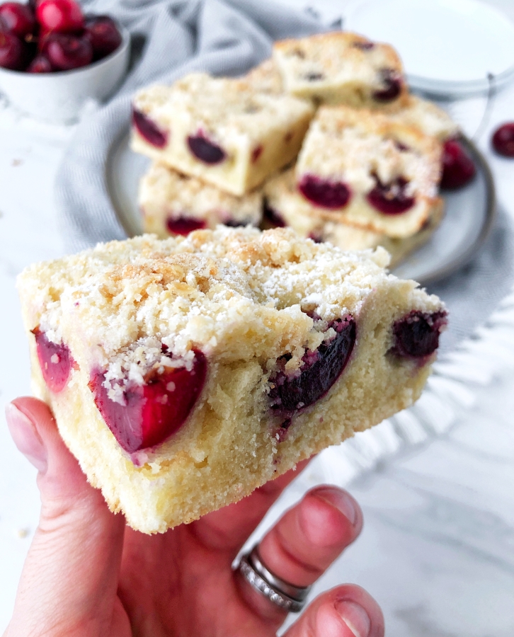 A hand holding cherry fruit cake, bars of the cake are on the plate in a background as well as cherries in a bowl